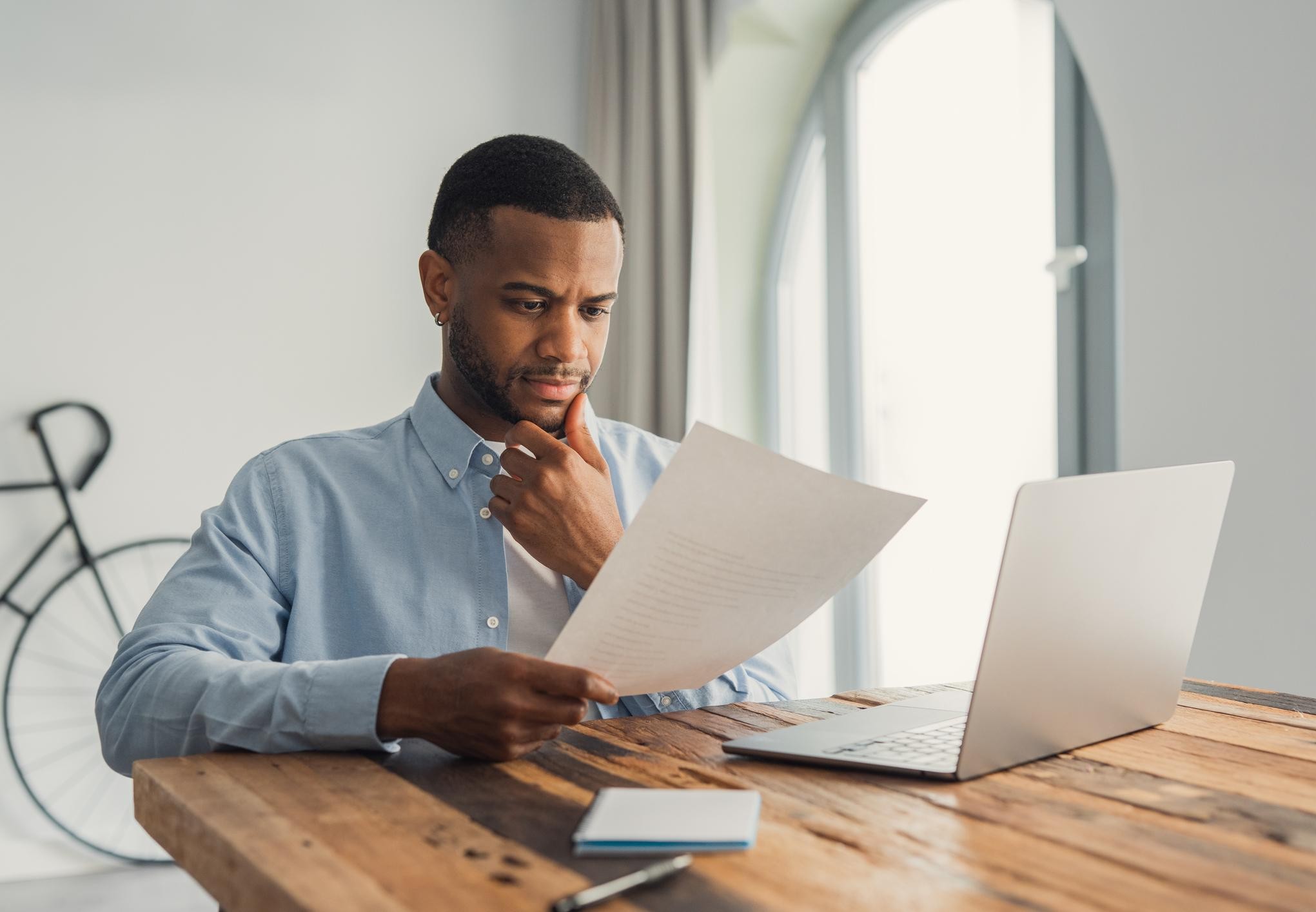 Man at a home office desk thoughtfully reviewing a job offer letter next to an open laptop